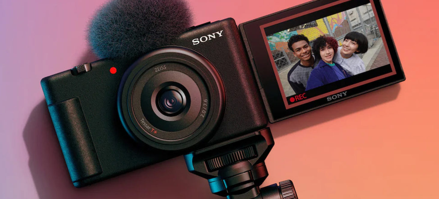 Una fotocamera nera Sony con microfono, su un supporto. Lo schermo mostra tre persone e la scritta "REC".