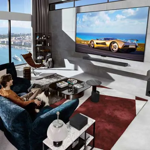 Soggiorno con divano blu, tavolino, tappeto rosso e grande TV con auto sportiva. Vista mare dalla finestra.
