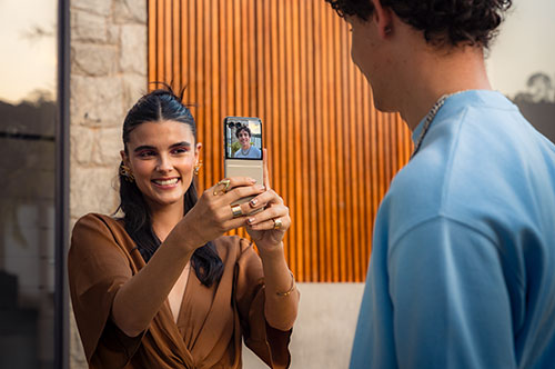 Una donna sorride mentre scatta una foto a un uomo con un telefono pieghevole. Uno sfondo a listelli di legno.