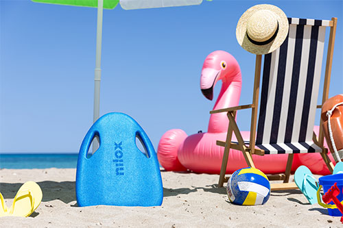 Spiaggia assolata con ombrellone, fenicottero gonfiabile rosa, sedia a sdraio a righe, tavola da surf blu e accessori da spiaggia.