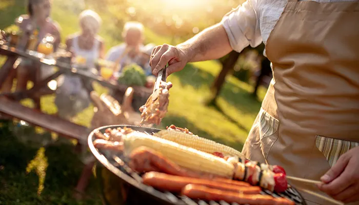 Grigliata all'aperto con mais, hot dog e spiedini su una griglia. Una persona con grembiule usa le pinze per girare il cibo. Persone sedute sullo sfondo.