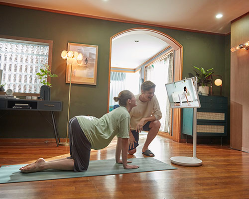 Donna su tappetino fa yoga, uomo guarda schermo. Finestra, scrivania, lampada e porta sullo sfondo.