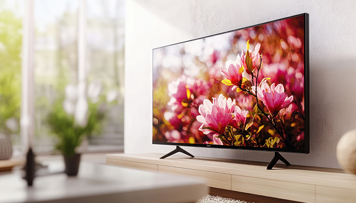 TV con schermo acceso mostra fiori rosa, posizionata su un mobile beige. Finestra luminosa sullo sfondo.
