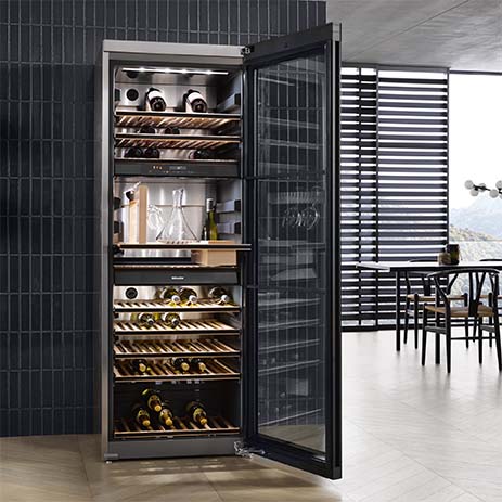 Frigorifero per vini aperto con ripiani in legno pieni di bottiglie. Tavolo e sedie in background.