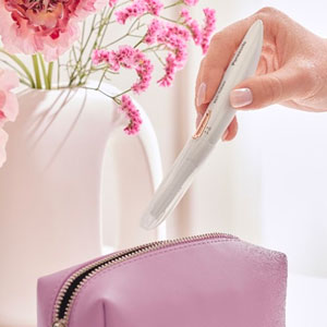 Una mano tiene uno strumento bianco sopra una borsa da toilette rosa. Dietro, fiori rosa in un vaso bianco.