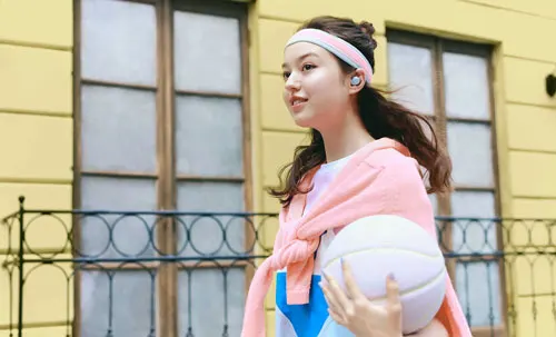 Ragazza con fascia e cuffie tiene un pallone da basket. Dietro, edificio giallo con finestre e ringhiera.