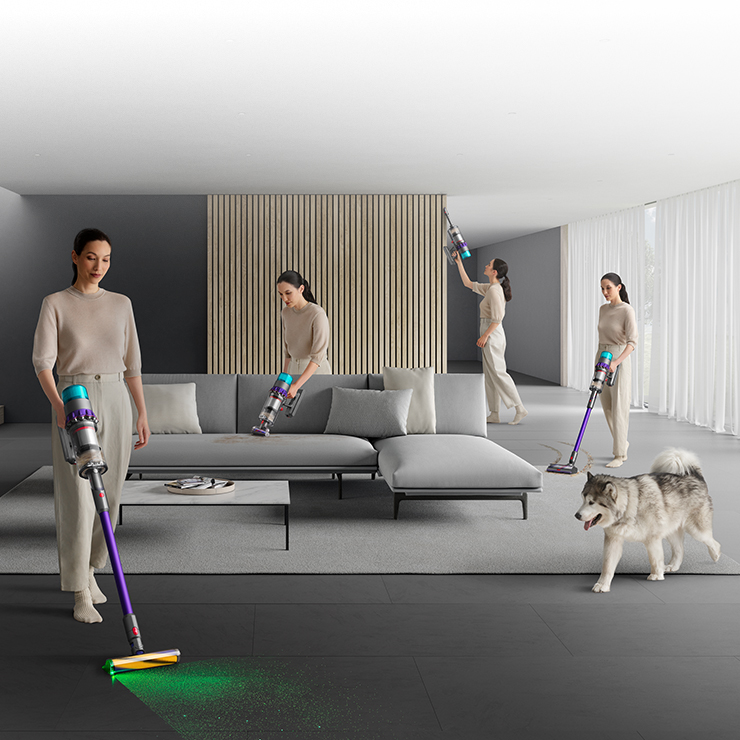 Interno di casa con donna che usa aspirapolvere viola e grigio, divano grigio, tavolino, cane e parete decorativa in legno.