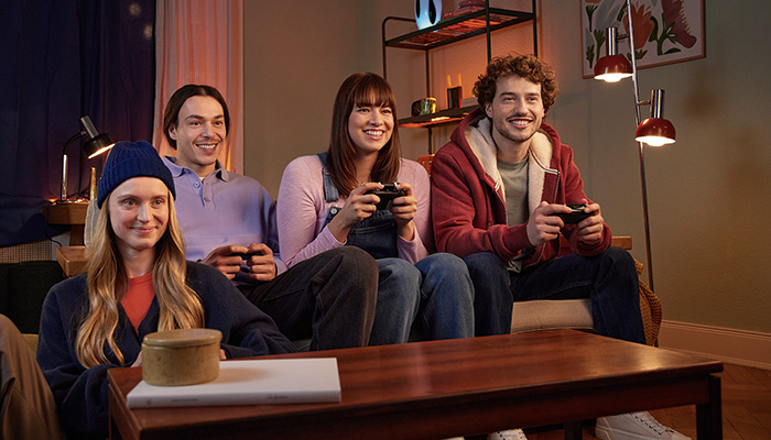 Quattro persone sedute su un divano giocano ai videogiochi con i controller. Un tavolino in legno è in primo piano.