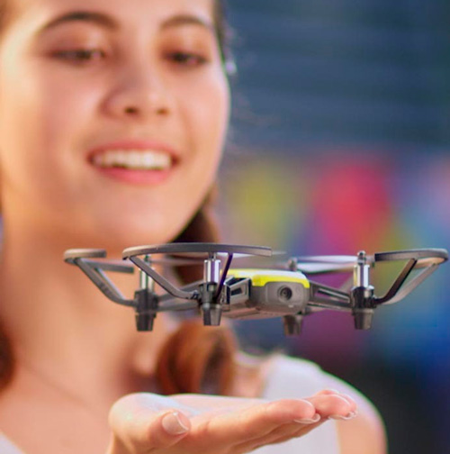 Donna sorridente tiene un drone grigio e giallo sul palmo della mano. Sfondo sfocato con colori vivaci.