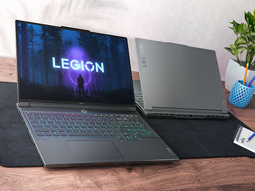 Due laptop grigi su scrivania di legno con tappetino nero. Uno aperto mostra logo "Legion" su schermo blu, l'altro � chiuso. Vaso con piante.