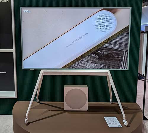 TV TCL su supporto bianco con rotelle, subwoofer quadrato. Sfondo verde scuro a righe verticali. Espositore marrone.