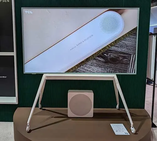 TV TCL su supporto bianco con rotelle, subwoofer quadrato. Sfondo verde scuro a righe verticali. Espositore marrone.