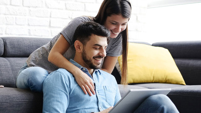 Uomo seduto su un divano grigio con tablet, donna in piedi dietro con le mani sulle sue spalle, cuscino giallo.