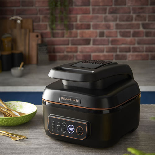 Friggitrice nera Russell Hobbs su un tavolo con ciotola verde e utensili dorati, su sfondo di mattoni.