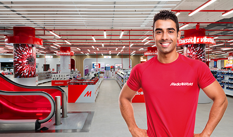 Uomo sorridente in maglietta rossa MediaWorld in negozio. Scale mobili rosse e scaffali di elettronica sullo sfondo.