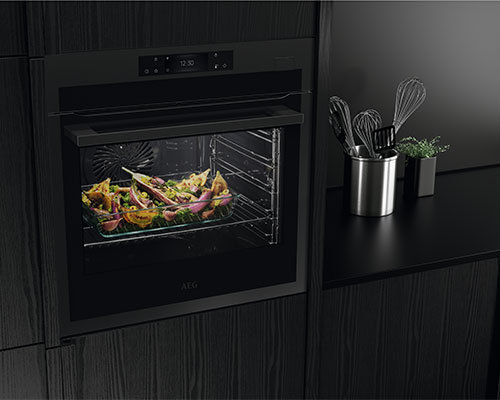 Forno nero con verdure all'interno. A destra, utensili da cucina in metallo e una pianta su un piano nero.