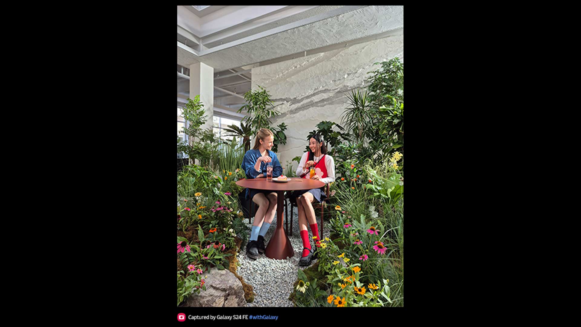 Due donne sedute a un tavolo rotondo in un giardino interno, tra piante verdi e fiori colorati.