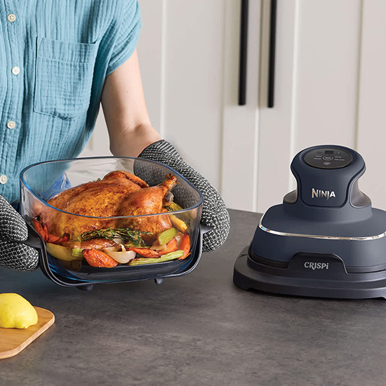 Una persona con guanti da forno lavorati a maglia regge un contenitore in vetro contenente un pollo arrosto intero e verdure, accanto alla friggitrice ad aria Ninja Crispi blu scuro.