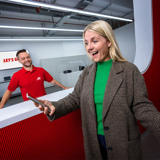 Donna sorridente con smartphone, indossa giacca a quadri e maglia verde. Dietro, uomo in polo rossa e scritta "Let's Go".