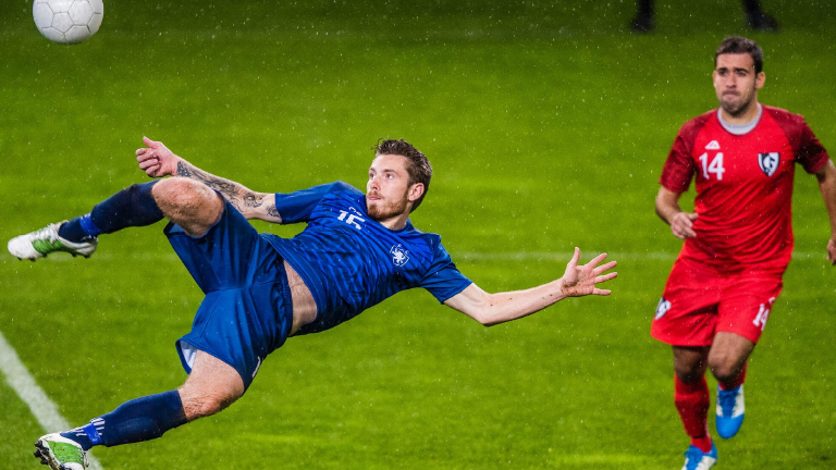 Un giocatore in blu calcia un pallone su un campo verde. Un altro giocatore in rosso corre sullo sfondo.