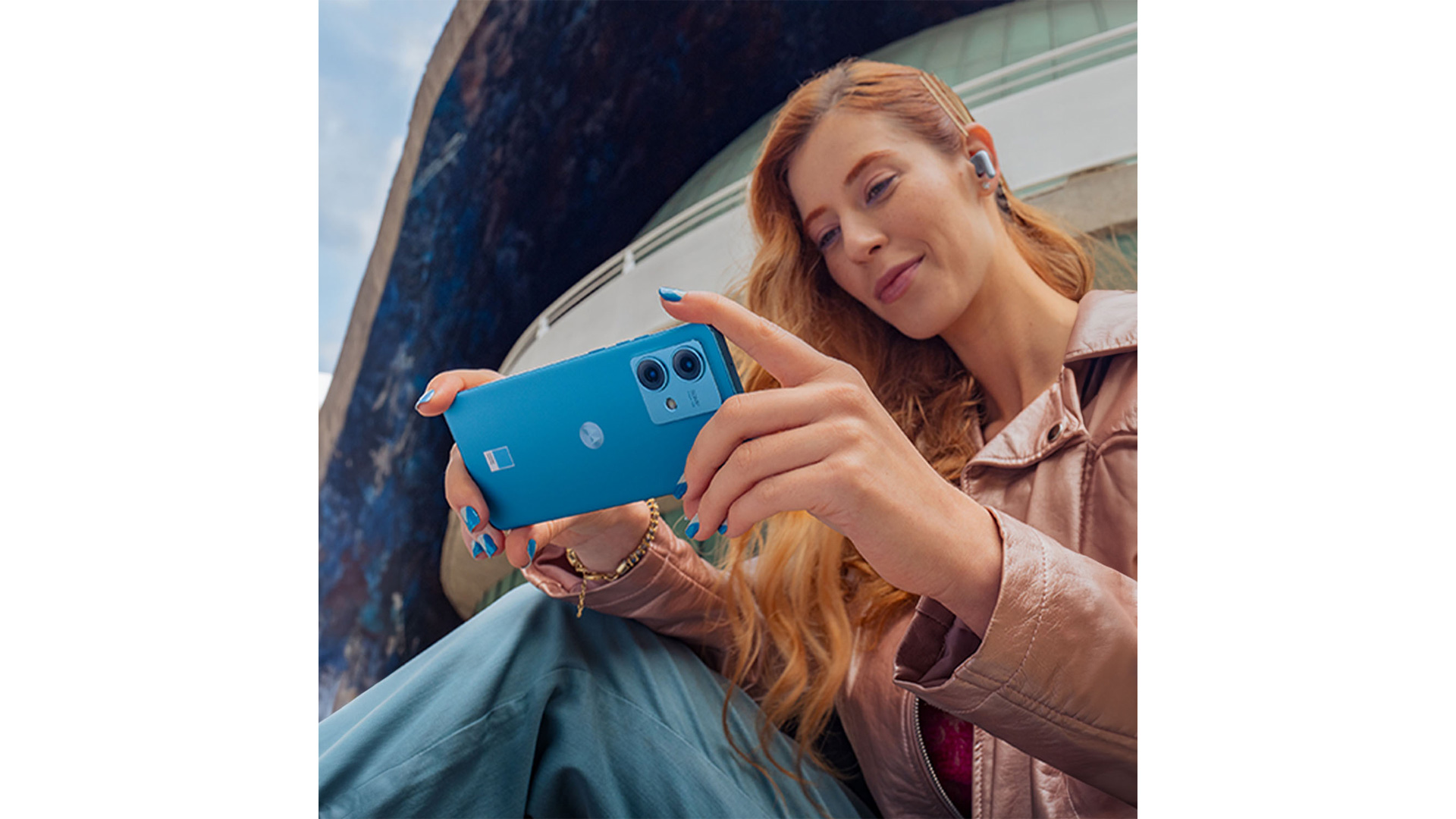 Donna con telefono azzurro, cuffie e giacca rosa. Sfondo architettonico.