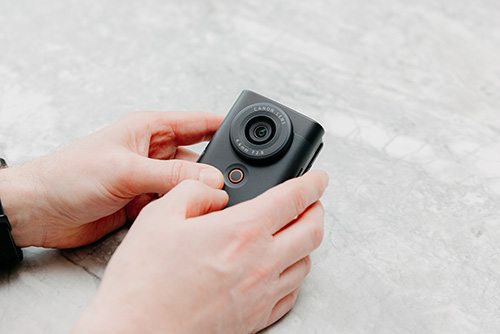 Una persona tiene in mano una fotocamera nera rettangolare con un obiettivo rotondo. Lo sfondo � una superficie grigia ruvida.