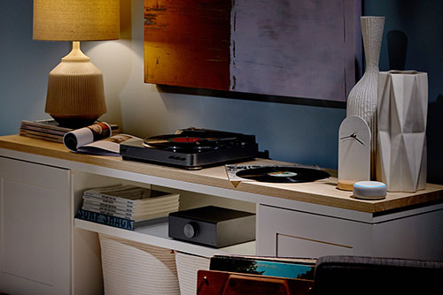 Mobile con giradischi nero, vaso bianco e lampada beige. Sopra, un disco in vinile e riviste. Sotto, altri oggetti e tessuti.