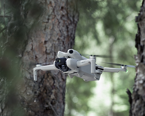 Drone bianco in volo tra alberi scuri. Lo sfondo � verde sfocato.