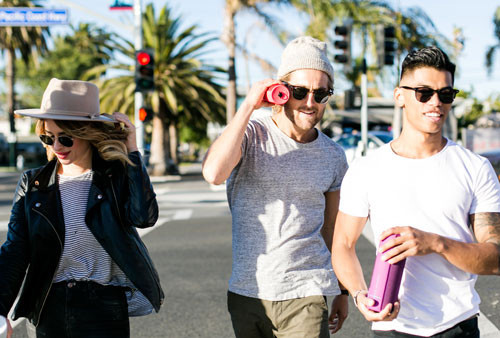 Tre persone attraversano la strada con palme e semaforo sullo sfondo. Indossano occhiali da sole.