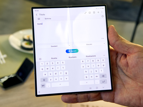 Mano che tiene un tablet aperto con tastiera touch. Sullo sfondo, un piatto con sushi e un telefono pieghevole.