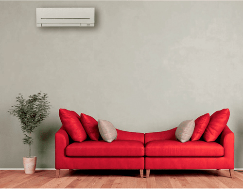 Soggiorno con divano rosso, cuscini, pianta in vaso e condizionatore su muro grigio chiaro, pavimento in legno.
