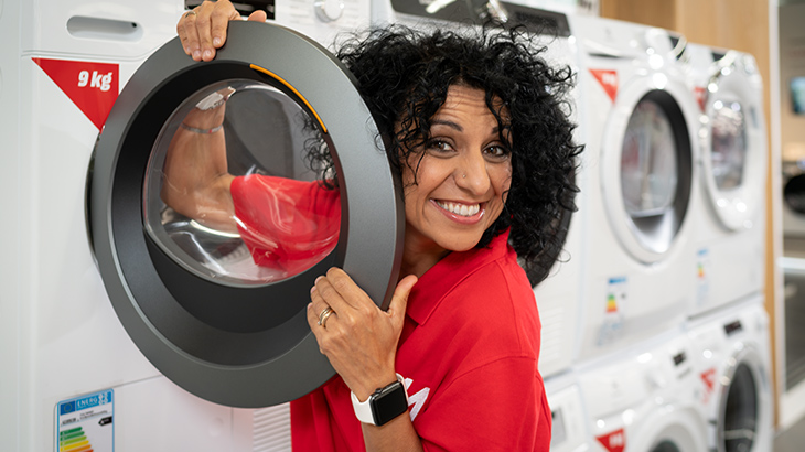 Donna sorridente in camicia rossa tiene l'obl� di una lavatrice bianca. All'interno, indumento rosso. Altre lavatrici sullo sfondo.