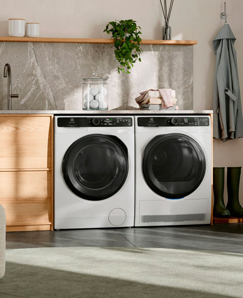 Lavanderia con lavatrice e asciugatrice bianche affiancate, mobile in legno chiaro e accessori decorativi. Stivali verdi a terra.