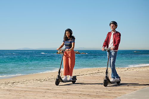 Due persone su monopattini su una spiaggia con elmetti. Dietro, l'oceano blu sotto un cielo azzurro.