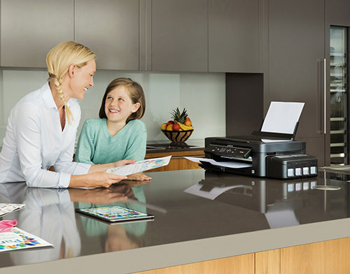 Donna e bambina sorridono in cucina. Stampante su ripiano vicino a frutta. Documenti e tablet sul piano.