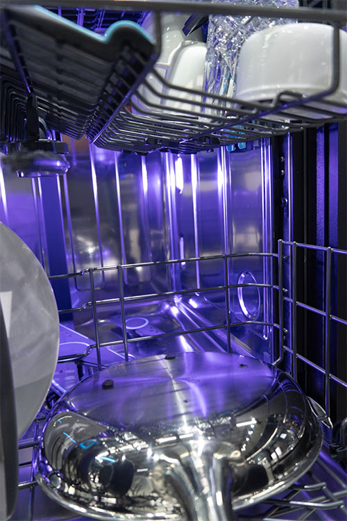 Interno di una lavastoviglie con piatti bianchi, bicchieri e una pentola d'acciaio, illuminata da una luce blu.