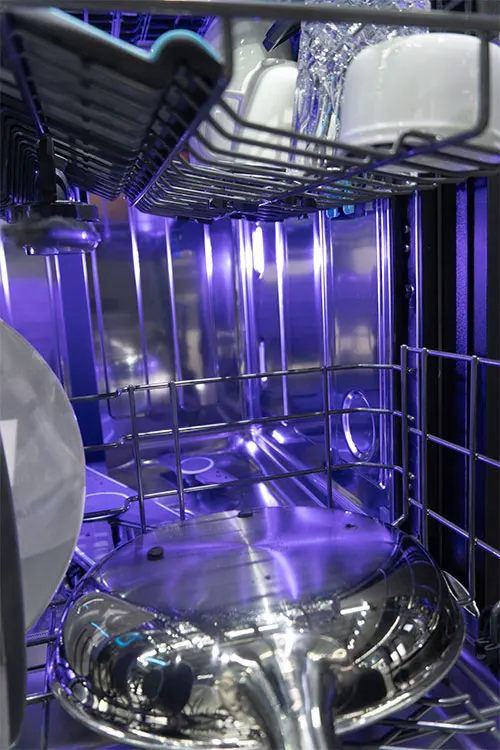 Interno di una lavastoviglie con piatti bianchi, bicchieri e una pentola d'acciaio, illuminata da una luce blu.
