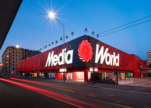 Edificio rosso e nero "MediaWorld" con insegna luminosa bianca, illuminato da lampione sotto cielo sereno. Tracce rosse di auto in strada.
