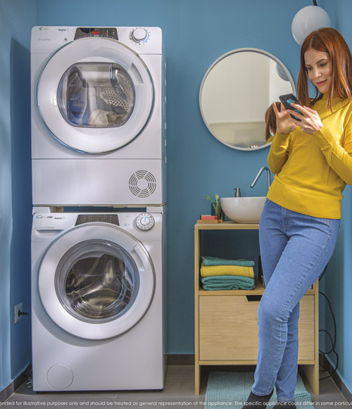 Donna in bagno con lavatrice e asciugatrice impilate, mobile lavabo con asciugamani e specchio rotondo, usa il cellulare.