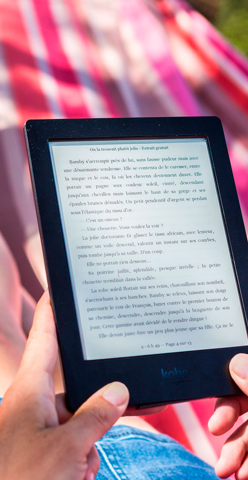 E-reader nero in mani, testo francese sullo schermo. Sfondo sfocato a righe rosa e bianche. Jeans e prato visibili.