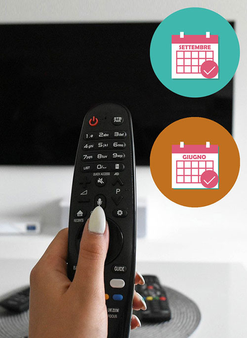 Primo piano di una mano che tiene un telecomando nero con icone di calendario in alto a destra. Sullo sfondo, una TV.