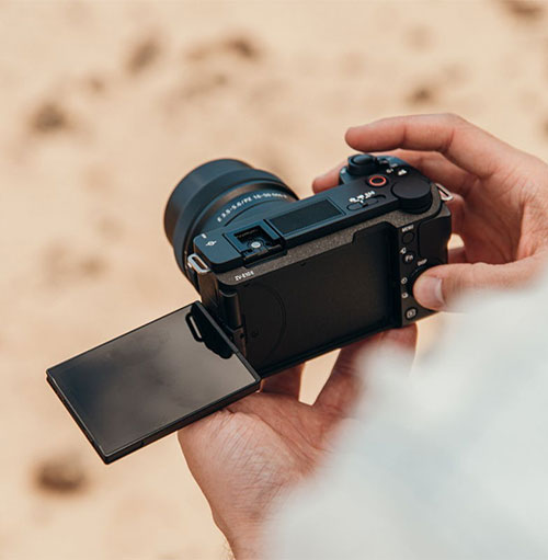 Una mano tiene una fotocamera nera con lo schermo aperto. Lo sfondo è beige chiaro, forse sabbia.