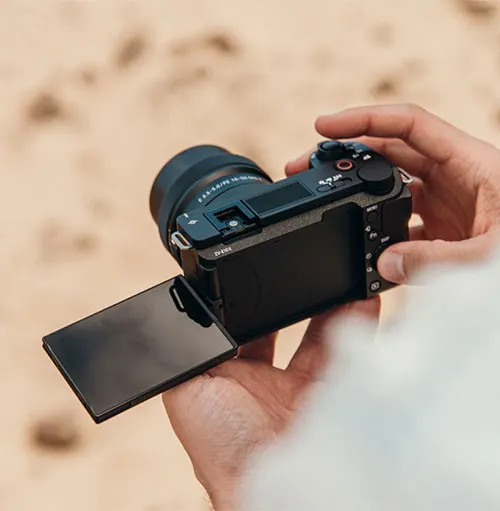 Una mano tiene una fotocamera nera con lo schermo aperto. Lo sfondo è beige chiaro, forse sabbia.