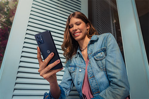 Donna sorridente con giacca di jeans che tiene un telefono nero davanti a una parete grigia a listelli.