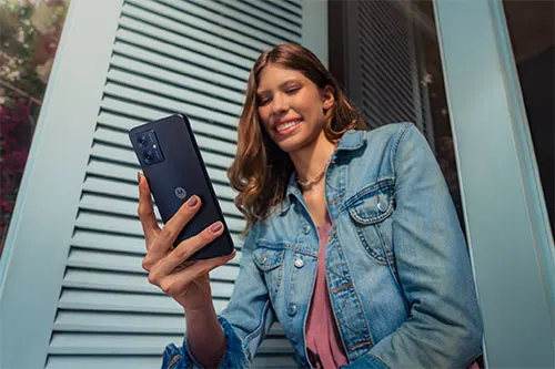 Donna sorridente con giacca di jeans che tiene un telefono nero davanti a una parete grigia a listelli.