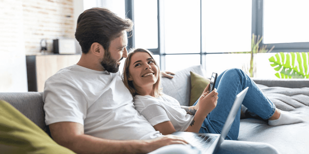 Uomo con laptop e donna con telefono su divano grigio. Finestra sullo sfondo e cuscini verdi.