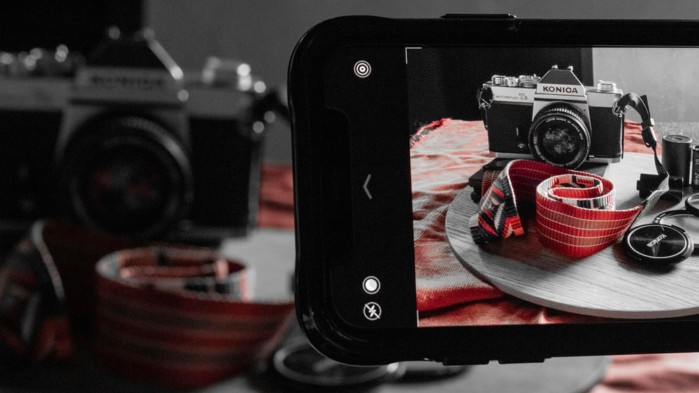 In un display si vede una Konica su un piatto, con accessori e una tracolla rossa. Un'altra fotocamera � sfocata sullo sfondo.