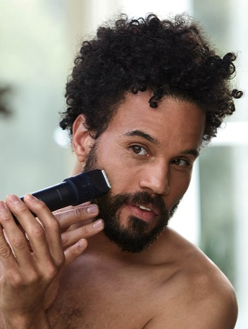 Uomo a torso nudo che si rade la barba con un rasoio elettrico nero, su sfondo sfocato chiaro.