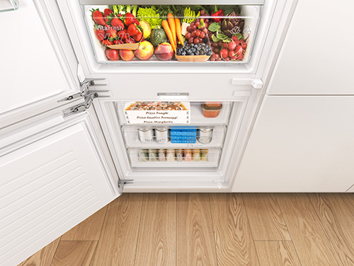 Vista di un frigorifero bianco aperto, pieno di frutta, verdura e contenitori alimentari su ripiani. Il pavimento � in legno chiaro.