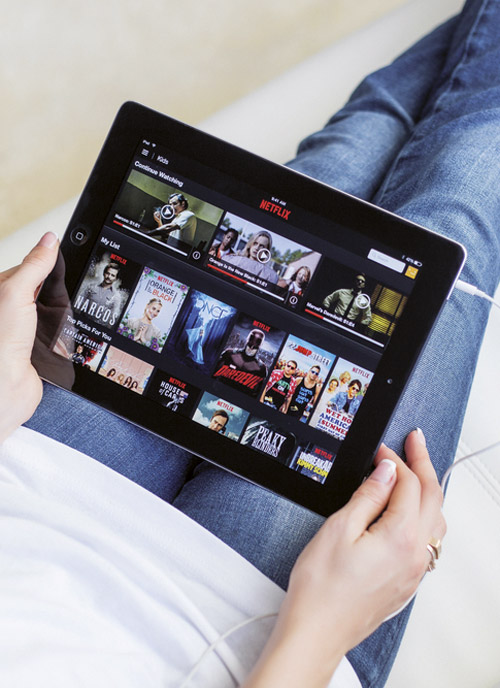 Una persona tiene un tablet con Netflix sulle gambe. Lo schermo mostra varie serie TV.